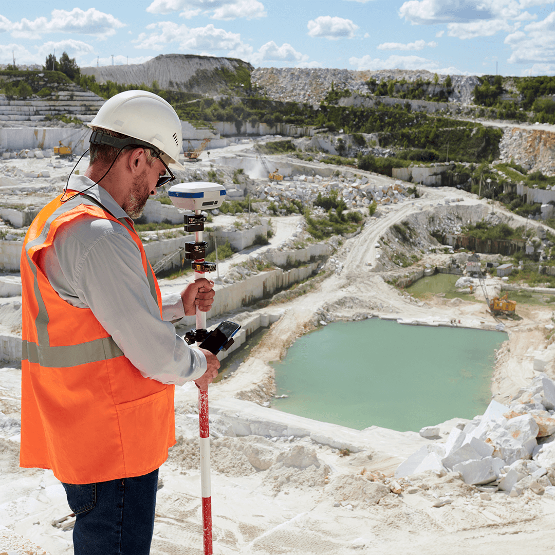 A surveyor in a high-visibility vest and hard hat uses GPS equipment in a large quarry with terraced rock, machinery, and a turquoise pond below.